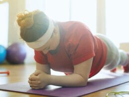 The woman, clad in a maroon T-shirt, is engaged in a plank position while lying down on a yoga mat