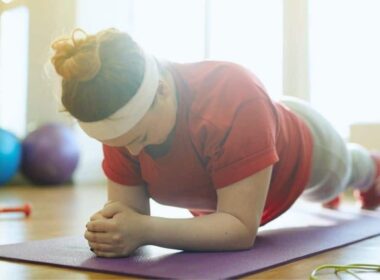 The woman, clad in a maroon T-shirt, is engaged in a plank position while lying down on a yoga mat