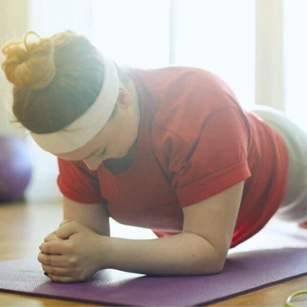 The woman, clad in a maroon T-shirt, is engaged in a plank position while lying down on a yoga mat